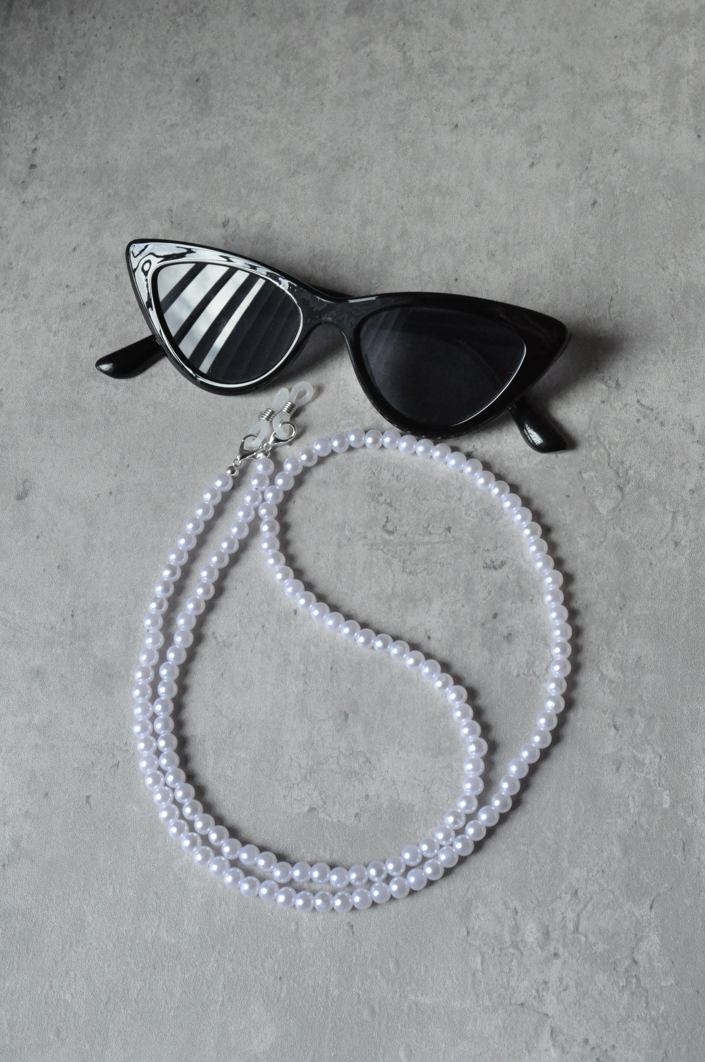 On a grey background, a pair of black cat-eye glasses is displayed with the Faux Pearl Glasses Holder draped over them. The chain features elegant pearls and a sophisticated design.