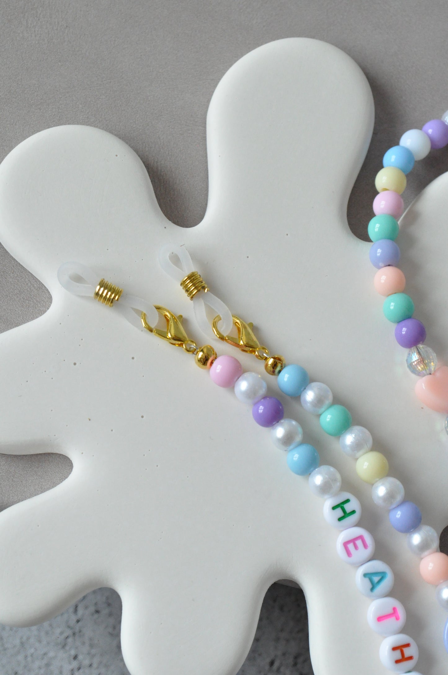 On a grey background, a close-up picture focuses on the gold-plated clasps and rubber holders of the Cute Pastel Beaded Glasses Chain, highlighting their high-quality craftsmanship.