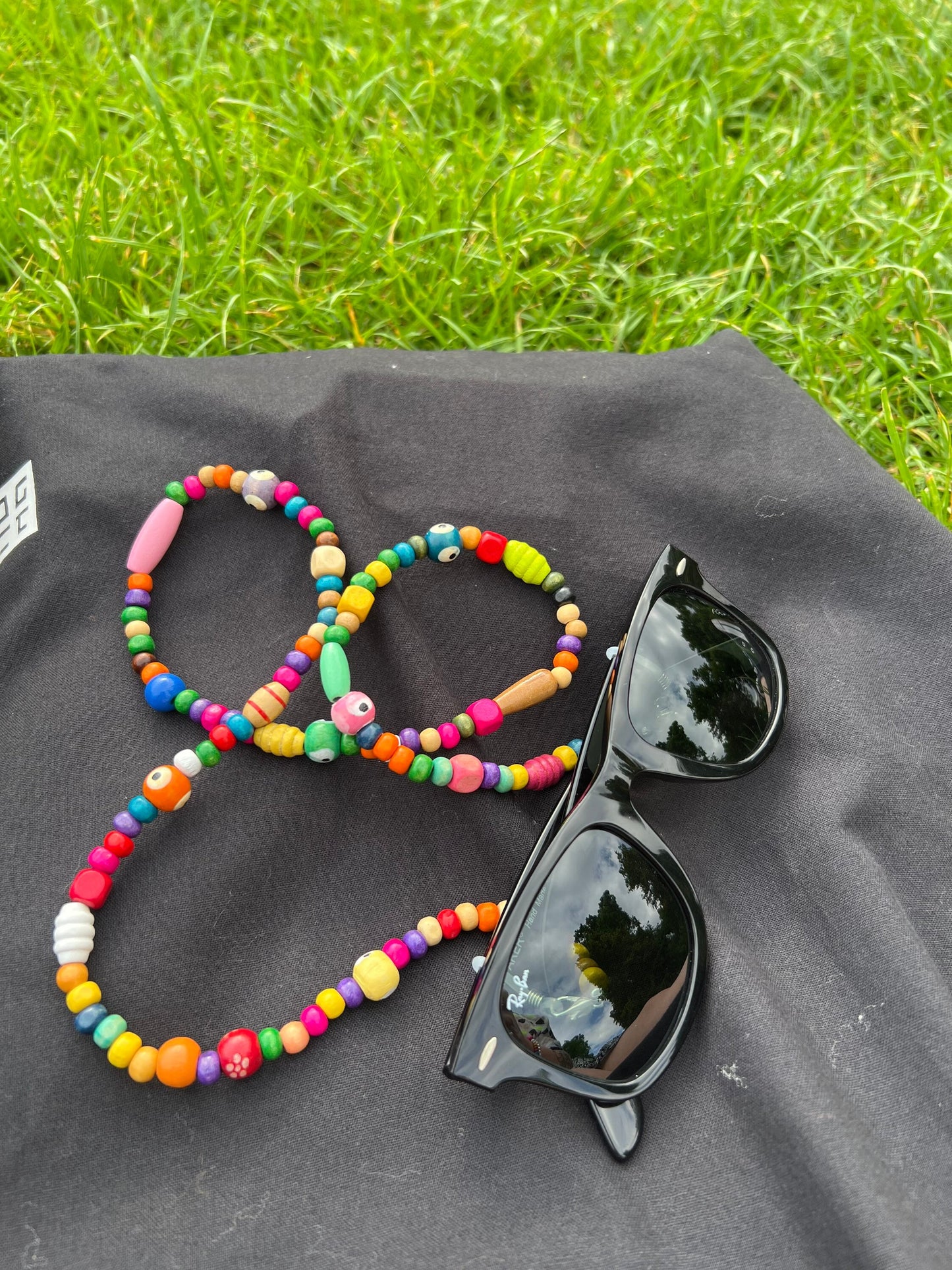 On a grassy surface, a black tote bag is positioned with black sunglasses and the Wooden Beaded Sunglasses Chain displayed on top. The chain features high-quality wooden beads and complements the sunglasses.