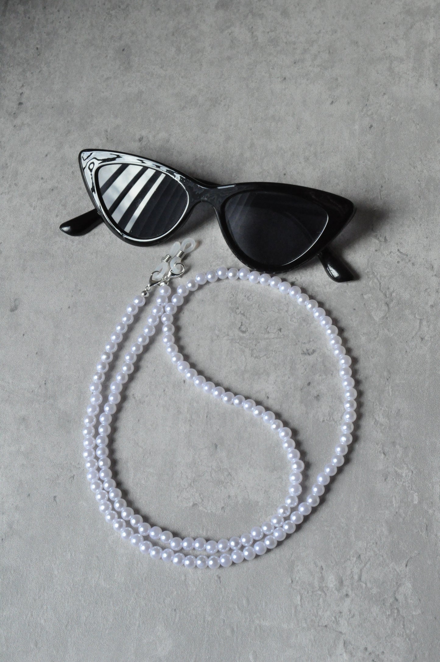  On a grey background, a pair of black cat-eye glasses is displayed with the Faux Pearl Glasses Holder draped over them. The chain features elegant pearls and a sophisticated design.