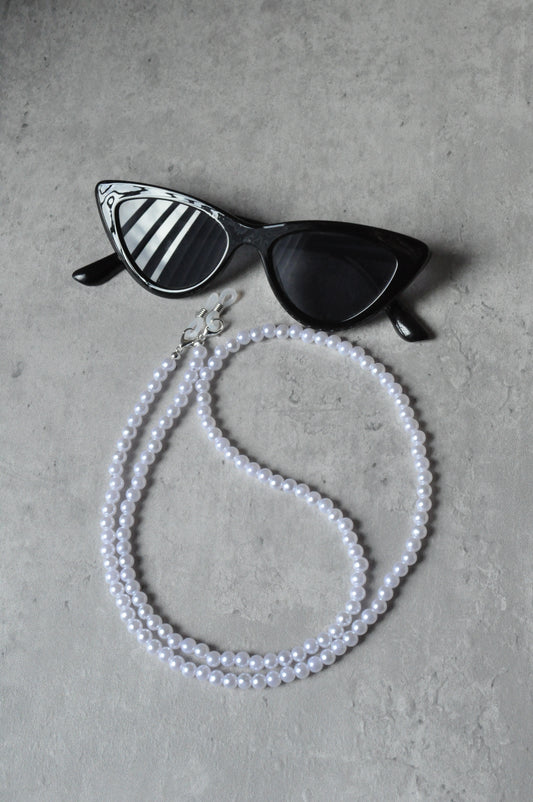  On a grey background, a pair of black cat-eye glasses is displayed with the Faux Pearl Glasses Holder draped over them. The chain features elegant pearls and a sophisticated design.