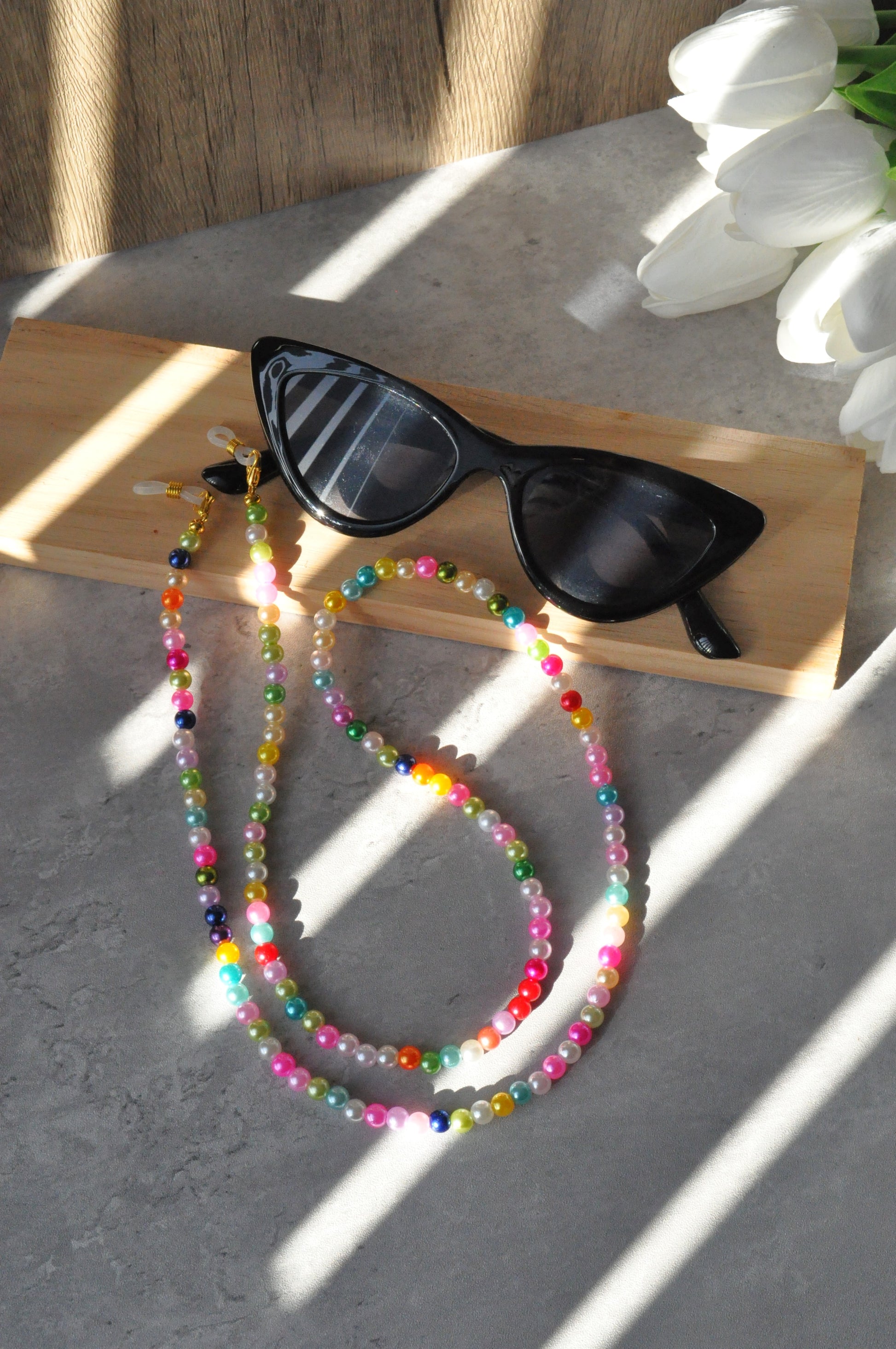 Cat eye glasses adorned with the Rainbow Colourful Beaded Glasses Chain, displayed on a small wooden piece with a white flower on the side, against a grey background.