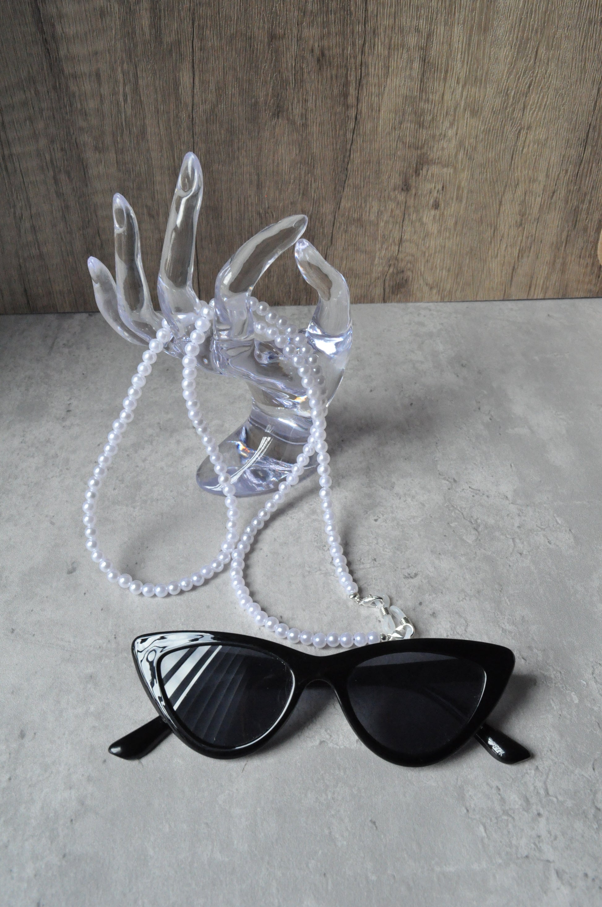 On a grey background, a plexiglass hand holds a Faux Pearl Glasses Chain, displayed alongside a pair of black sunglasses.