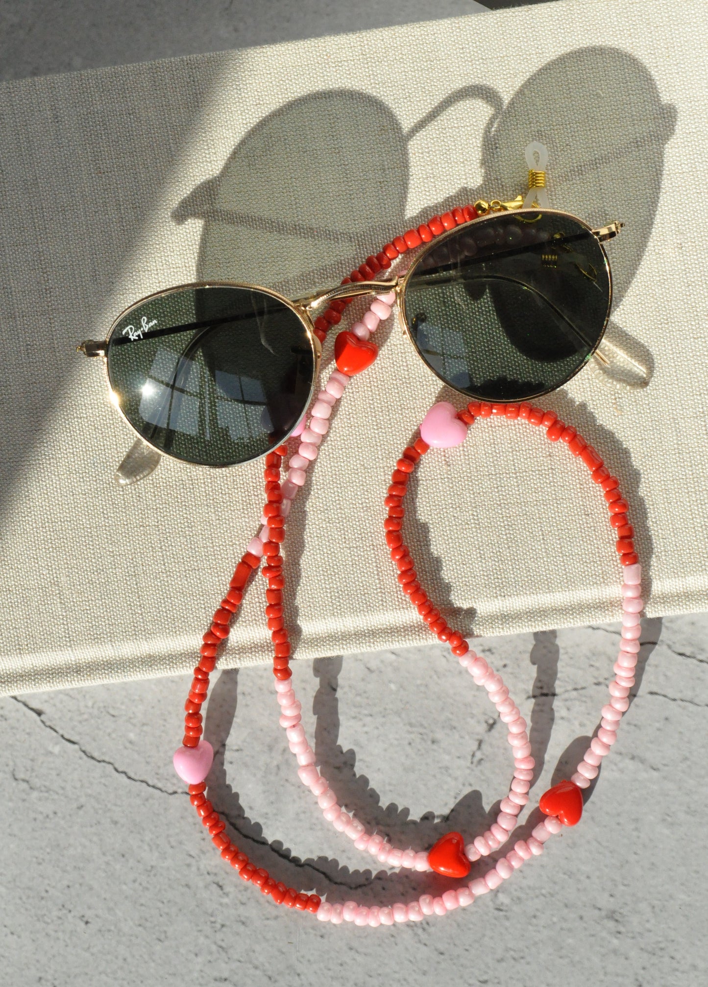 Pink and red hearts glasses chain displayed with round glasses on a textured surface against a grey background, shown from a different angle.