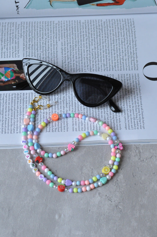 Fruit Salad Pastel Beaded Glasses Chain displayed with glasses on an open magazine, set against a grey background.