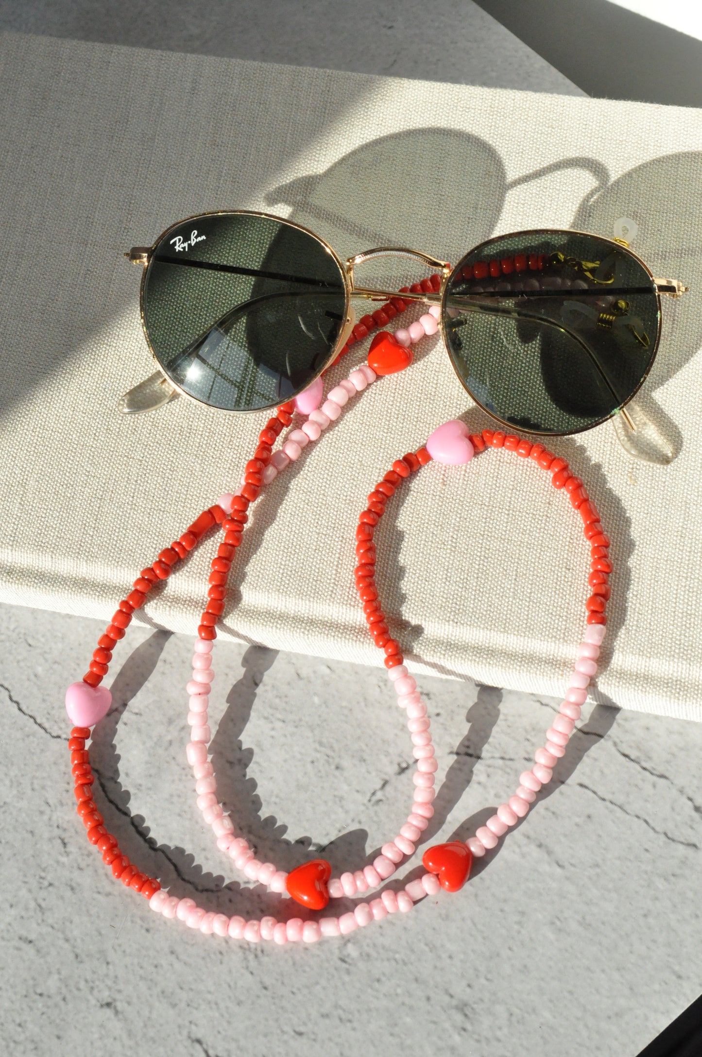 Pink and red hearts glasses chain displayed with round glasses on a textured surface against a grey background, shown from a different angle.