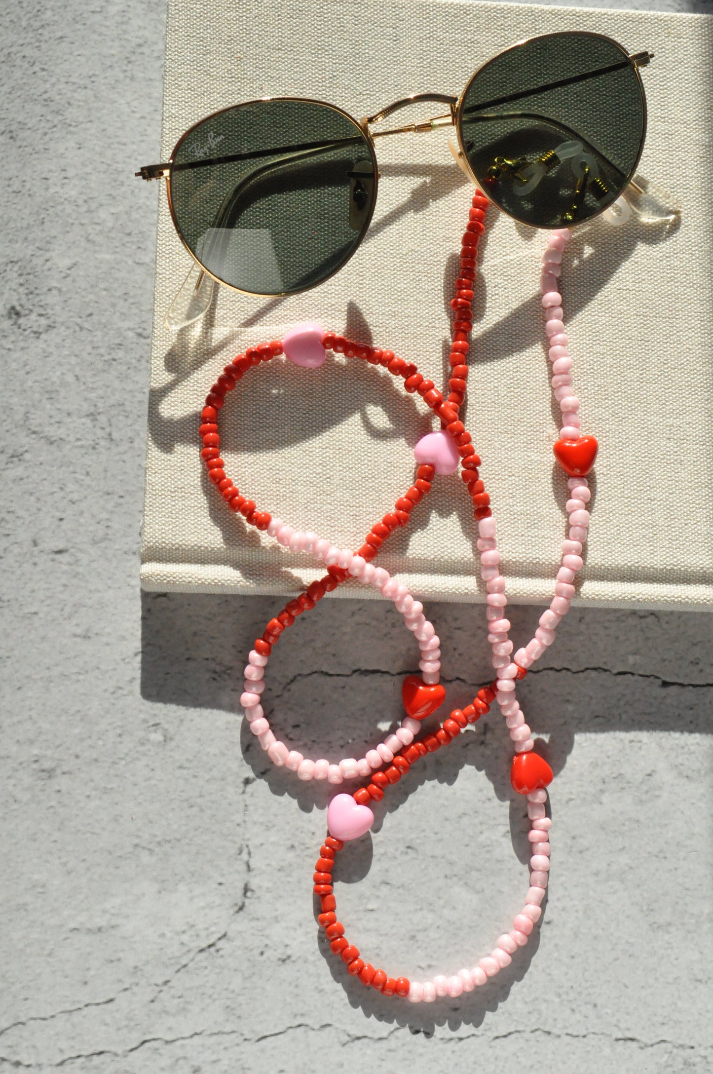 Pink and red hearts glasses chain displayed with round glasses on a textured surface against a grey background.