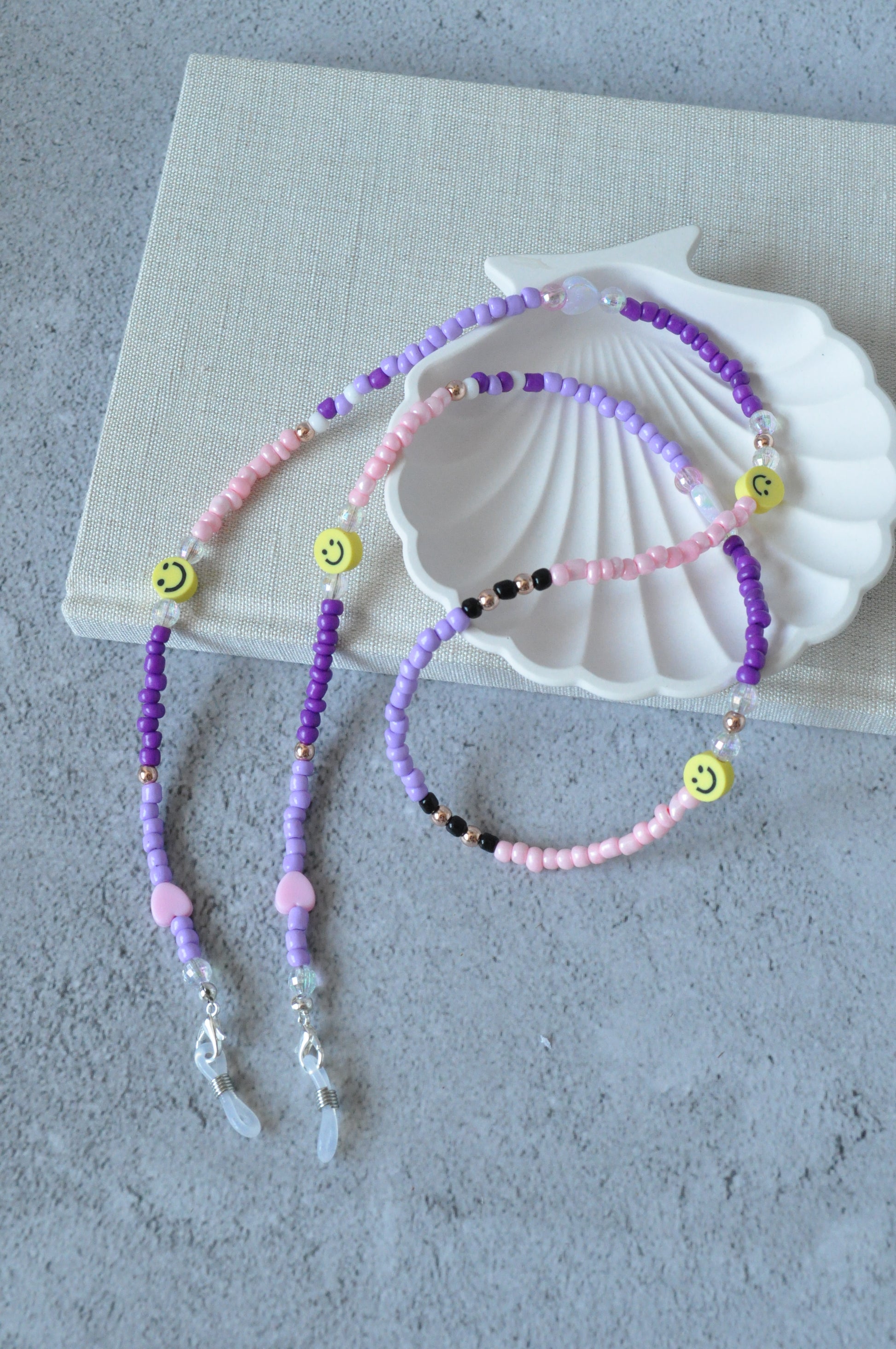  On a grey background, a textured notebook and a shell trinket dish are positioned. A lilac beaded glasses chain featuring smiley beads and rose gold accents is draped over the items.