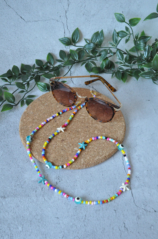 Colourful evil eye beaded glasses chain displayed on a cork surface against a grey background, with artificial leaves for added aesthetic.