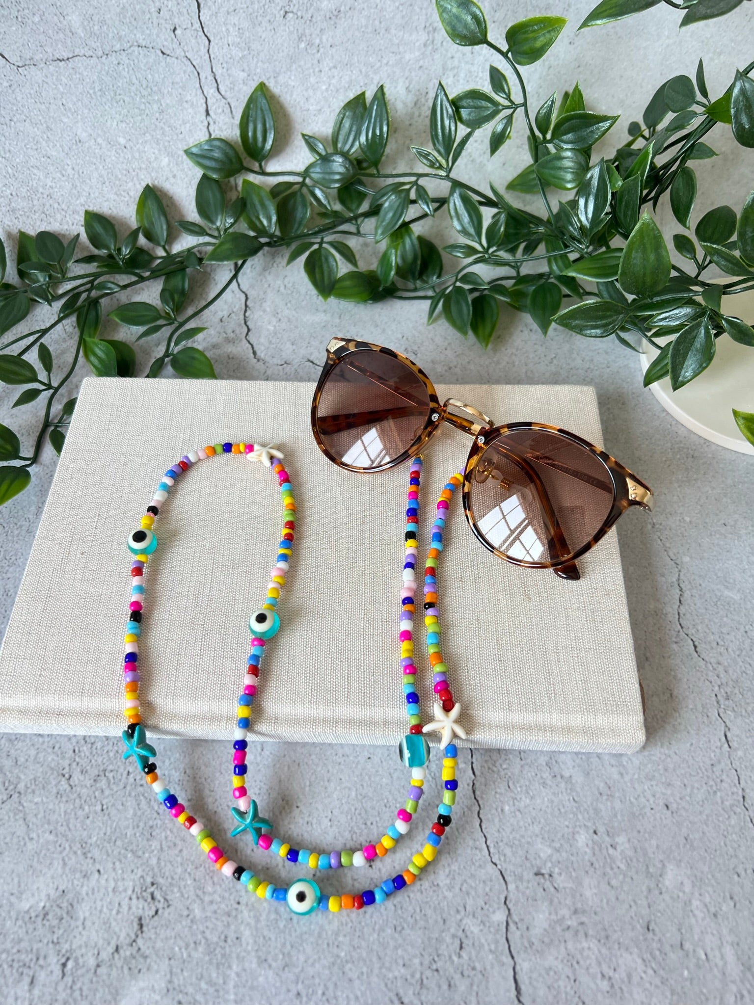 Colourful evil eye beaded glasses chain displayed on a cork surface against a grey background, with artificial leaves for added aesthetic.