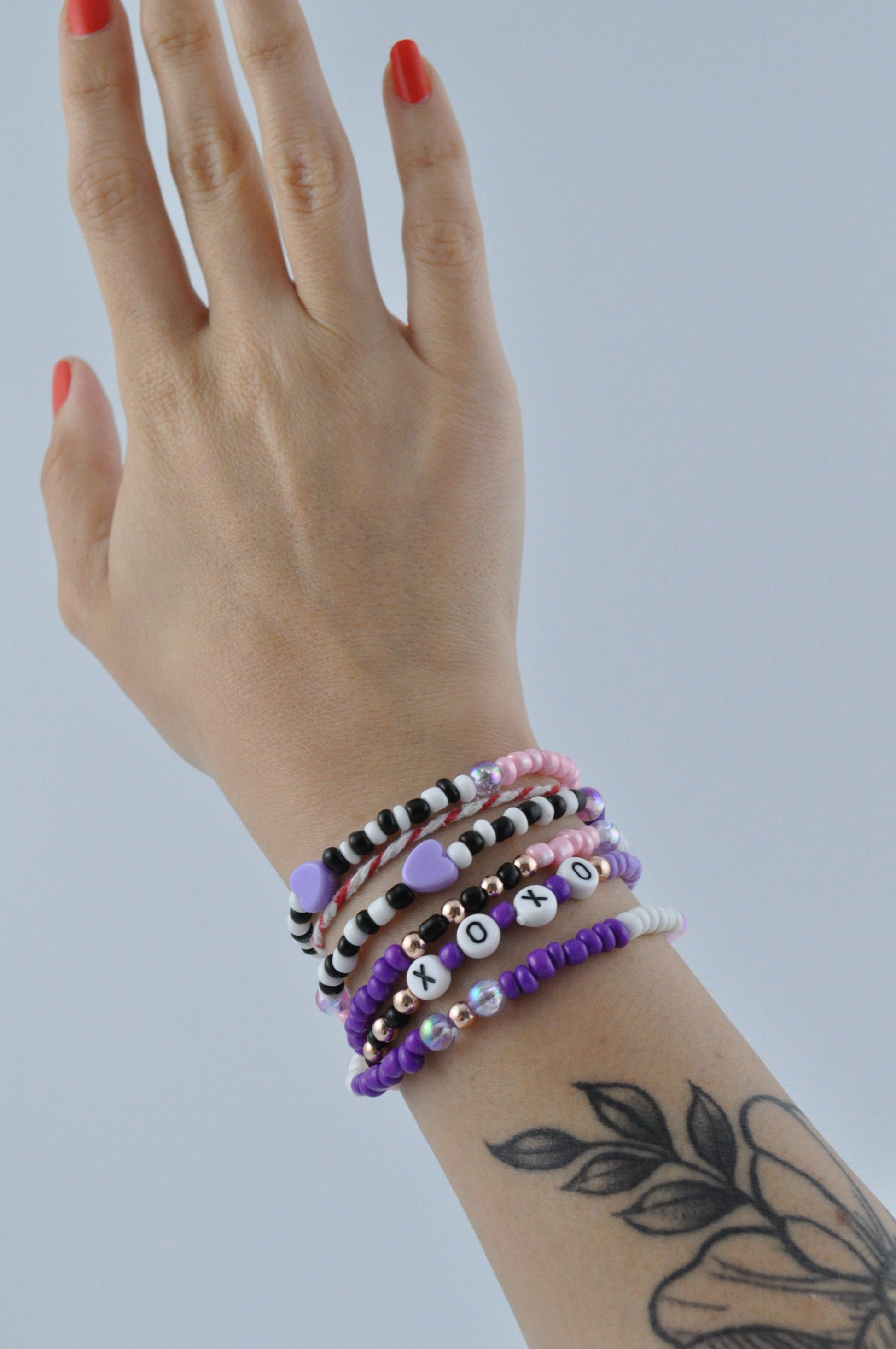 An arm with a flower tattoo and red nails showcases the lilac and rose gold beaded glasses chain worn as a layered bracelet. The chain is elegantly draped around the wrist, highlighting its vibrant colours and personalised "XOXO" detail.