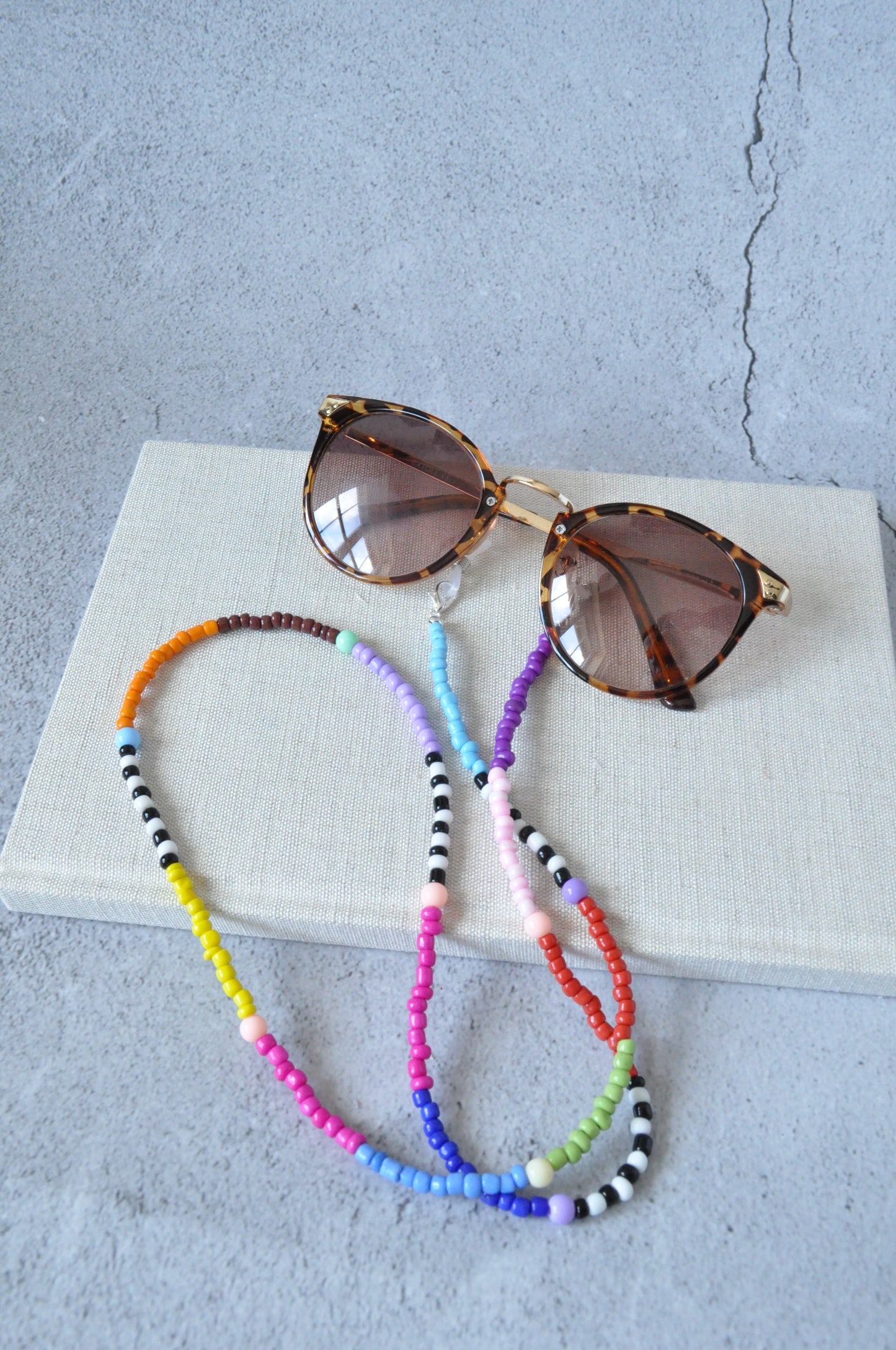 A vibrant rainbow beaded glasses chain is displayed on a grey background. A beige textured notebook sits at the top of the image, with a pair of leopard-print sunglasses adorned with the colourful glasses chain resting on it.