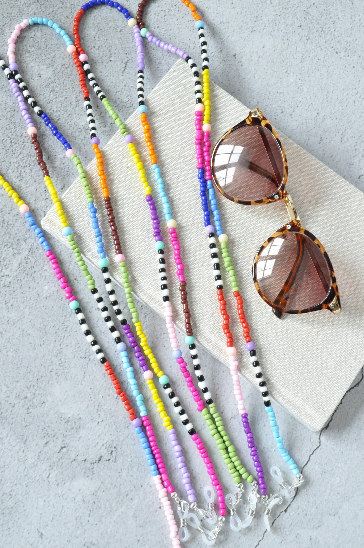 Four vibrant rainbow beaded glasses chains are displayed on a grey background. Each chain is arranged on a beige textured notebook, alongside a pair of leopard-print sunglasses adorned with the colourful chains.