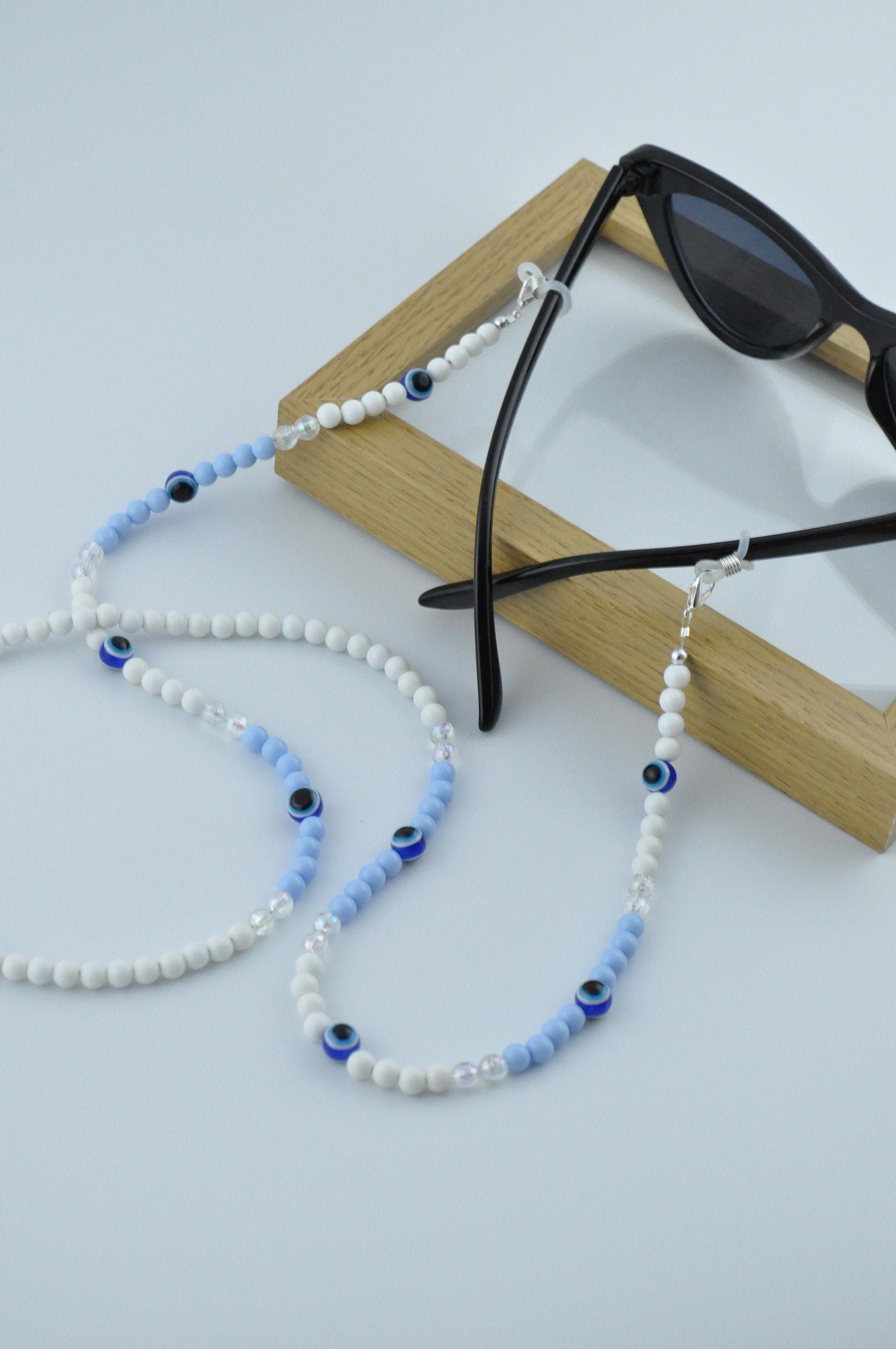 On a white background, a wooden frame is positioned with the blue Evil Eye Beaded Glasses Chain displayed on top. The chain features high-quality beads and the iconic Evil Eye symbol.