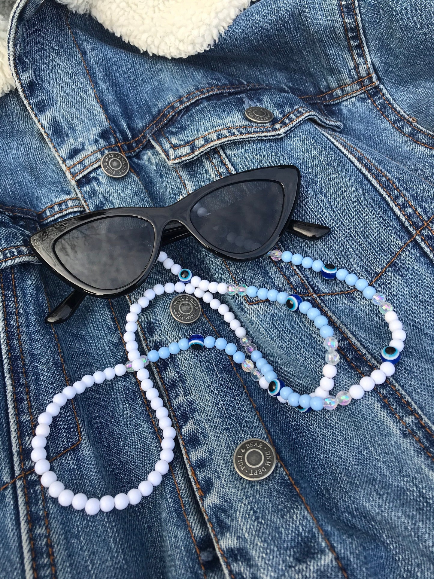 On a denim jacket, a pair of black cat-eye sunglasses is displayed with a blue Evil Eye Beaded Glasses Chain draped over them. The chain features high-quality beads and the iconic Evil Eye symbol.