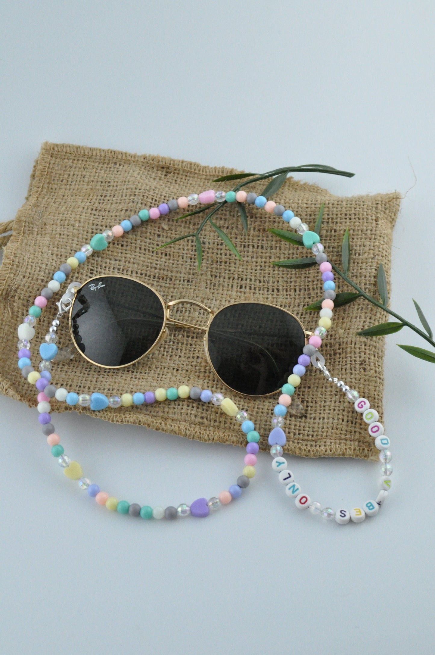 On a white background, a jute fabric is laid out with fake green branches arranged around it. Black round glasses are positioned on top of the fabric, showcasing the Cute Pastel Beaded Glasses Chain.