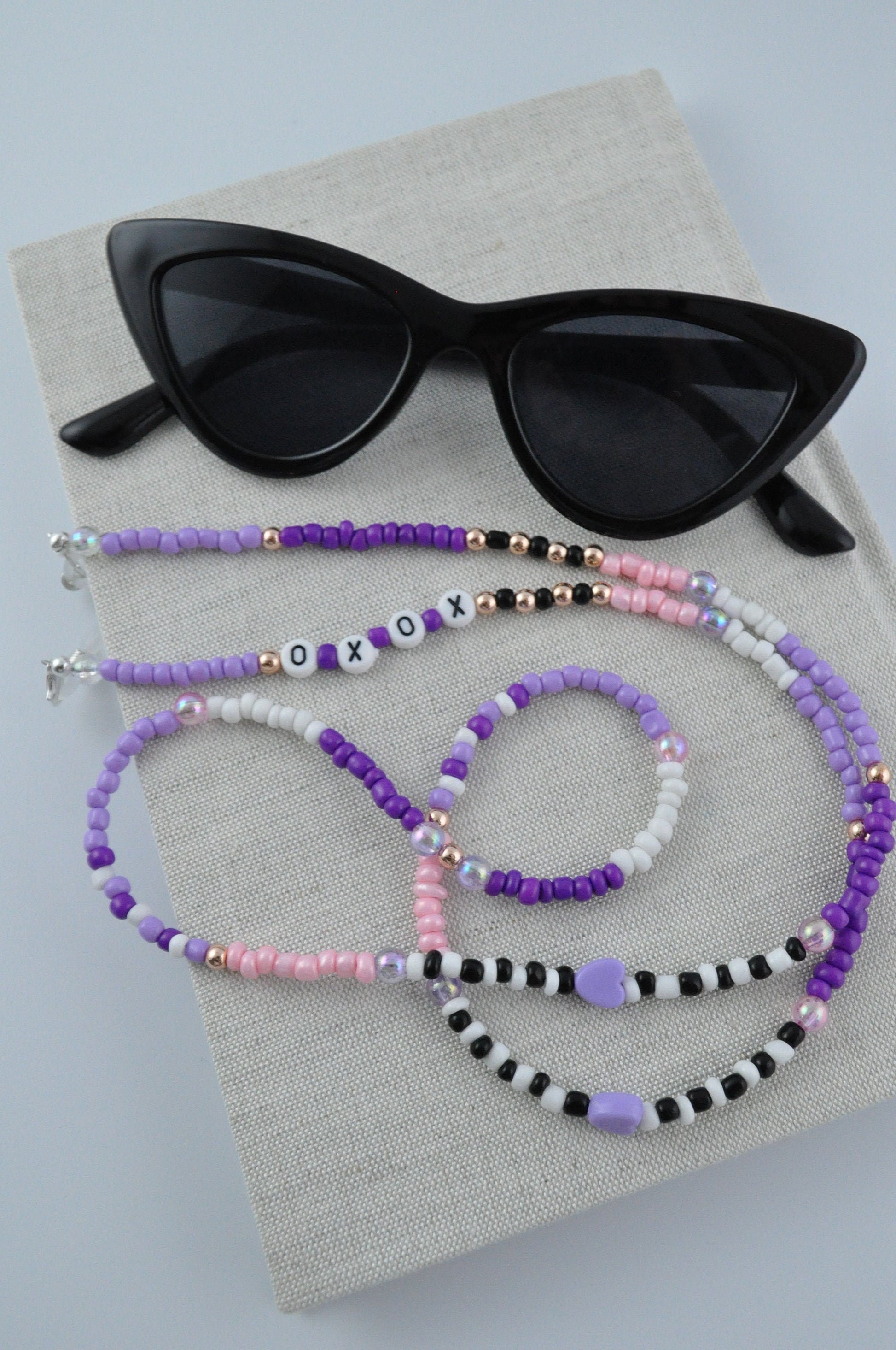  On a grey background, a textured notebook is displayed with a pair of black cat-eye sunglasses resting on it. The sunglasses are adorned with a lilac and rose gold beaded chain, personalised with "XOXO".